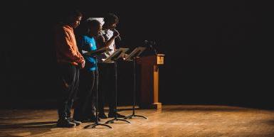 Three Black students, wearing casual clothing, stand to the right side of the image, facing toward the center of the image,behind black music stands on a warm wood stage. They are lit by a spotlight, with nearly all but themselves, part of the stage, and a wooden podium in shadow. 